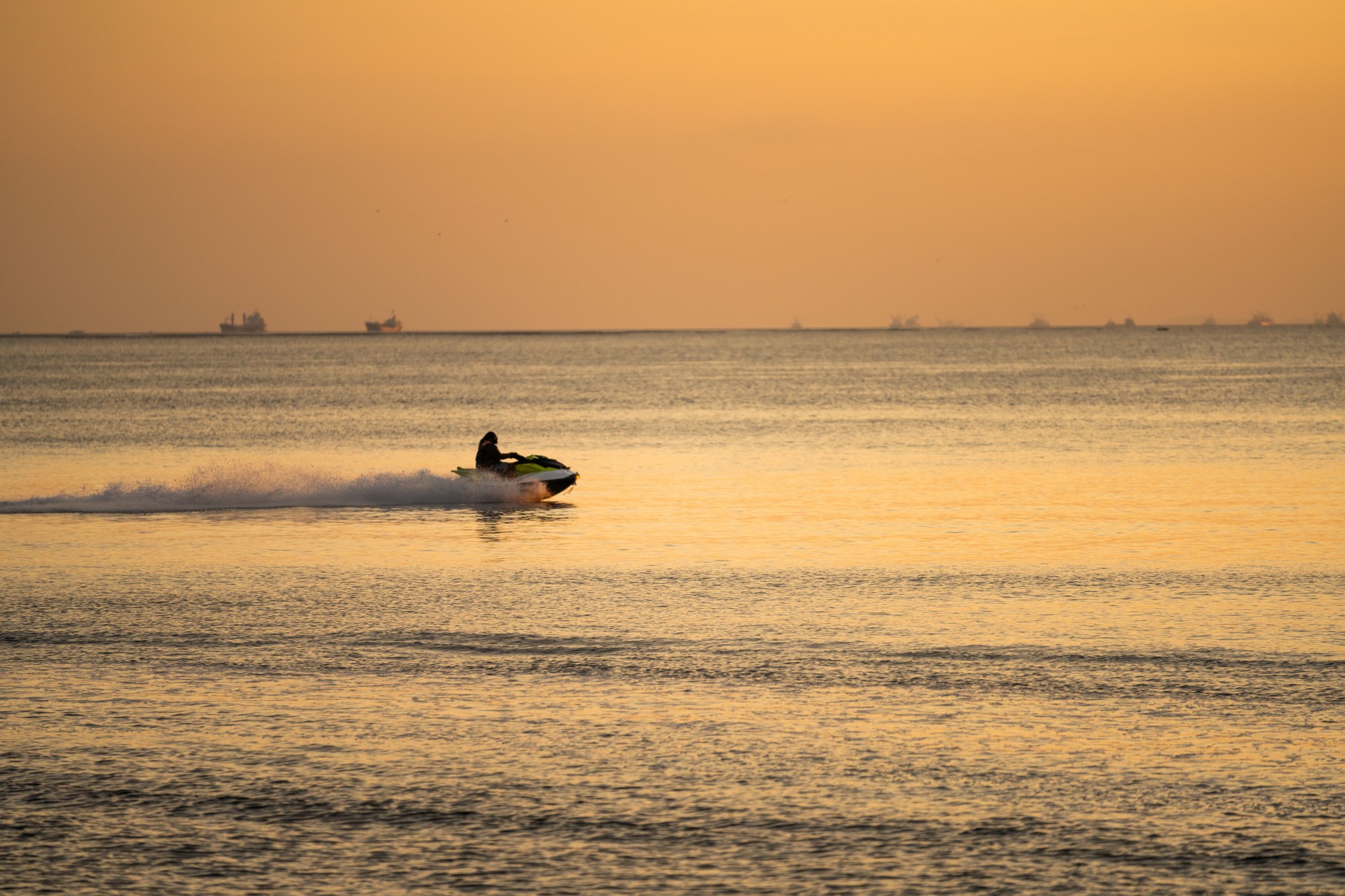 Jet ski rider enjoying a sunset on calm waters near the coast with ships in the background